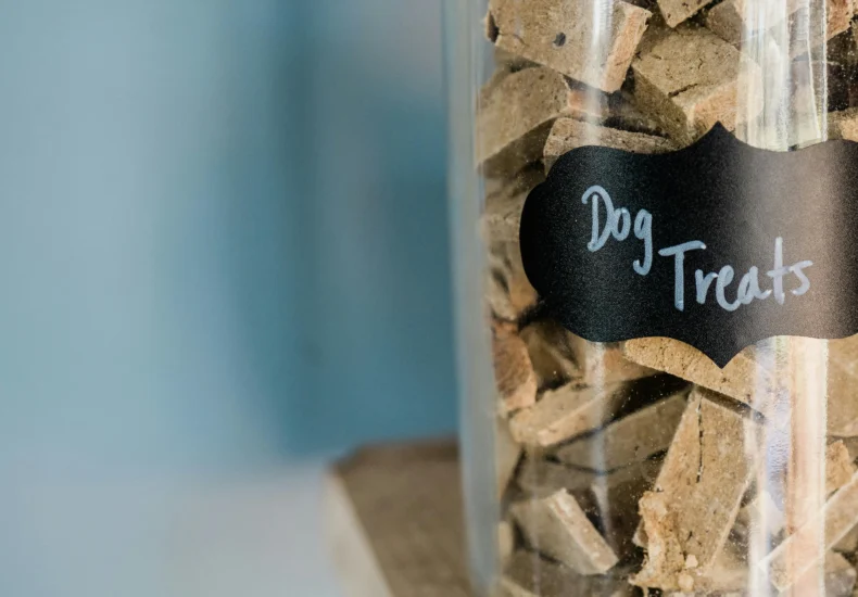 A close-up image of dry dog treats stored in a labeled glass jar on a wooden surface.