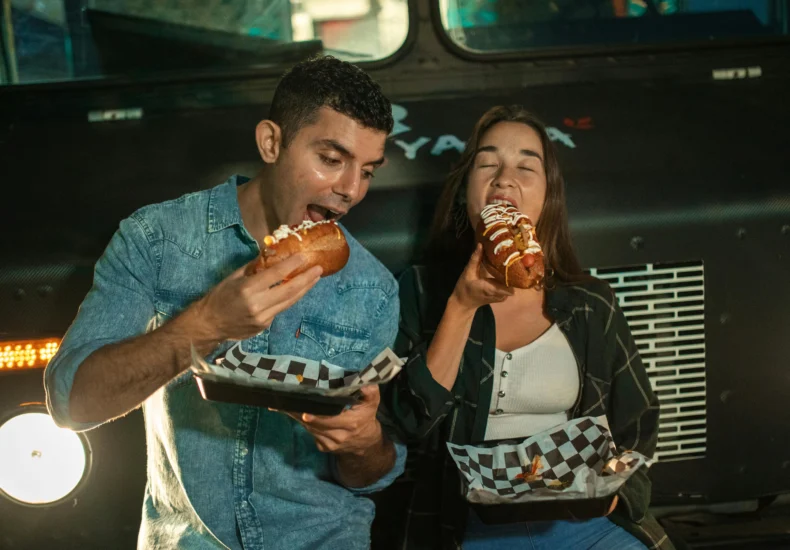 Happy couple eating hot dogs at a food truck, enjoying a casual night out.