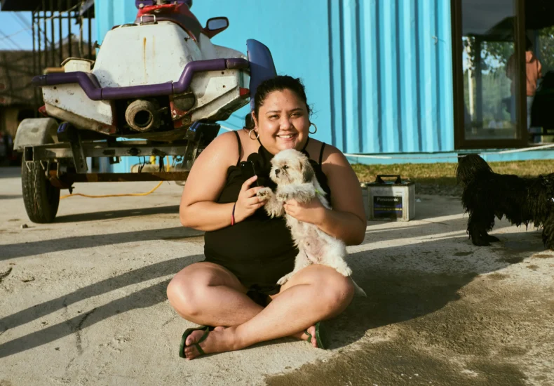 A woman in a black tank top holds two dogs, sitting in front of a colorful building.