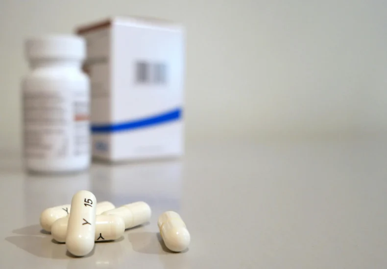 Close-up image of white capsules with medicine bottle and box in the background.