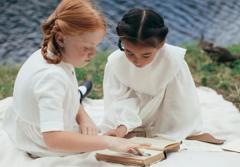 Two young girls sitting by a tranquil lakeside, enjoying a classic book on a summer day.
