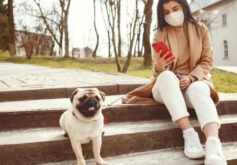Woman wearing mask sits on steps with pug, using a smartphone. Outdoors, casual and relaxed.