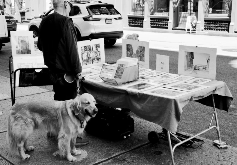 Black and white photo of a street vendor with artwork and dog in an urban setting.