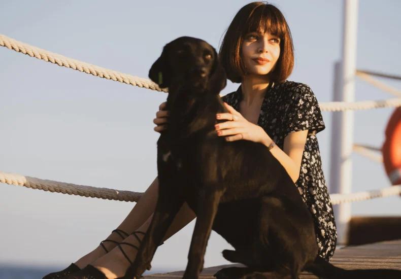 A young woman enjoys a sunny day sitting on a pier with her pet dog.