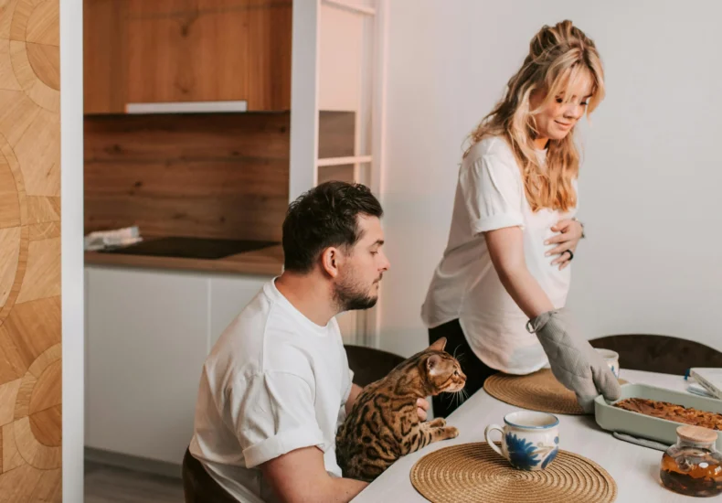 Expectant mother serves dinner while partner and cat sit nearby in a cozy dining space.