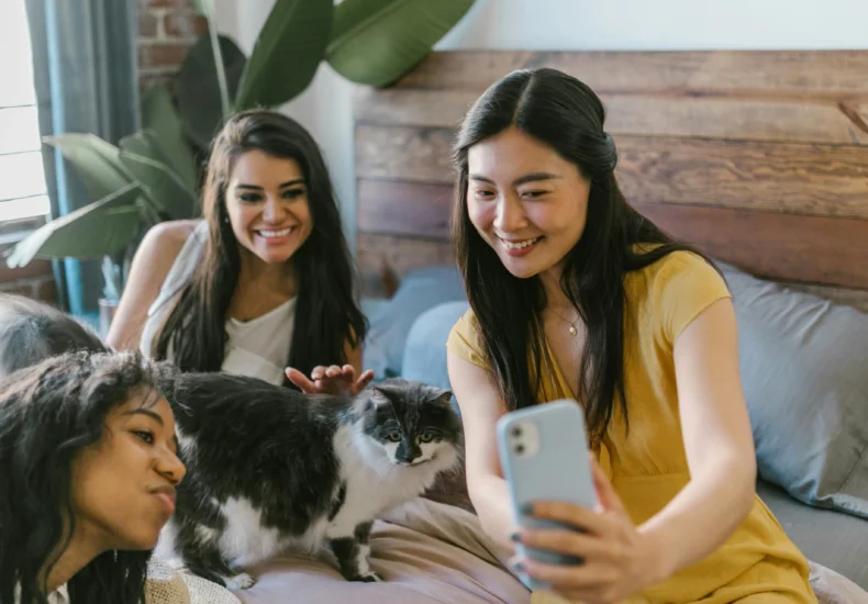 Three women smiling and taking a selfie with a cat in a cozy bedroom setting.