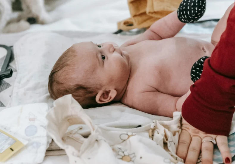 A mother changes her baby's diaper on the bed while a pet dog watches attentively.