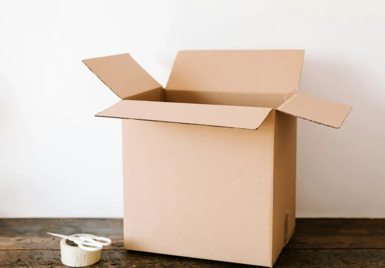 Opened cardboard container placed on old wooden desk near tape and scissors before relocation against white wall