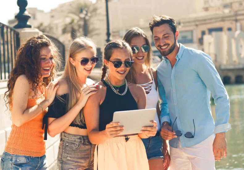 Five friends laughing together by the waterfront in Malta, enjoying a sunny day and sharing a tablet.