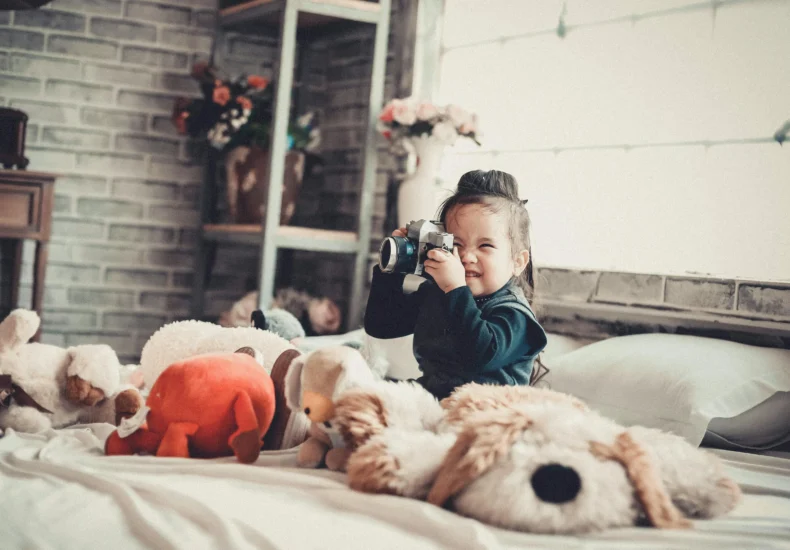 Smiling child with a camera surrounded by stuffed toys in a cozy bedroom setting.