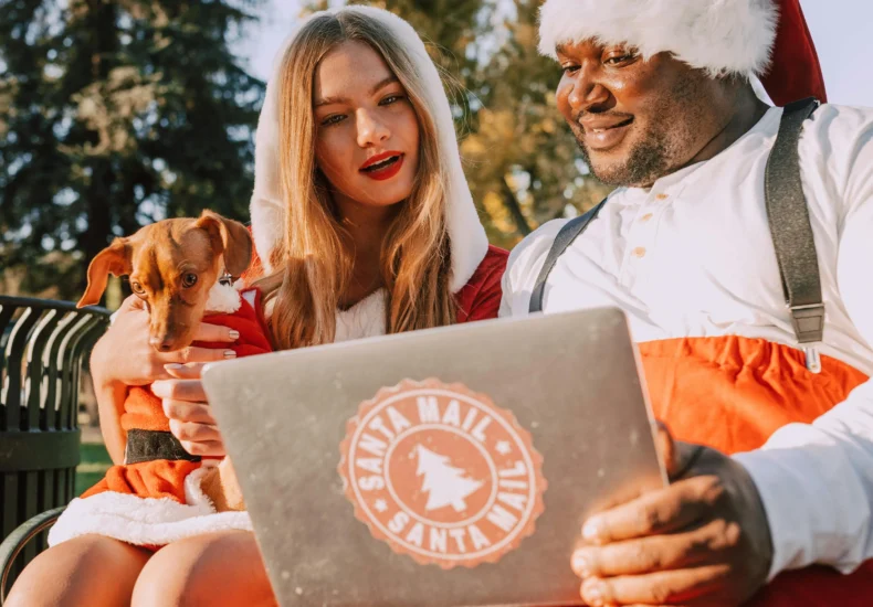 African American man and Caucasian woman in Santa outfits with laptop and dog outdoors.