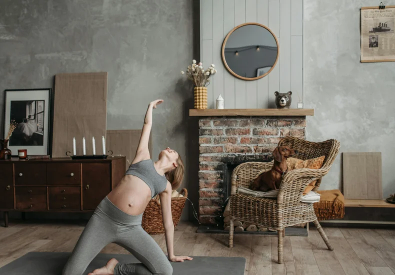Pregnant woman practicing yoga near a fireplace with a dog in a cozy room.