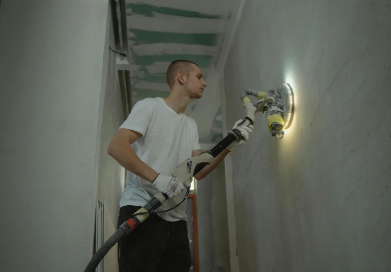 A man sanding an interior wall with an automatic sanding tool during renovation work indoors.