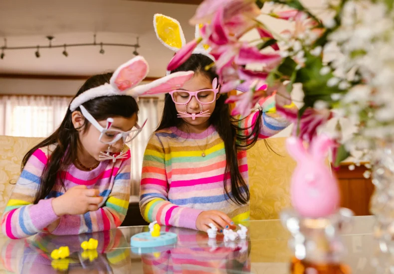 Two girls celebrate Easter indoors, wearing bunny ears and playing with toys at a table.