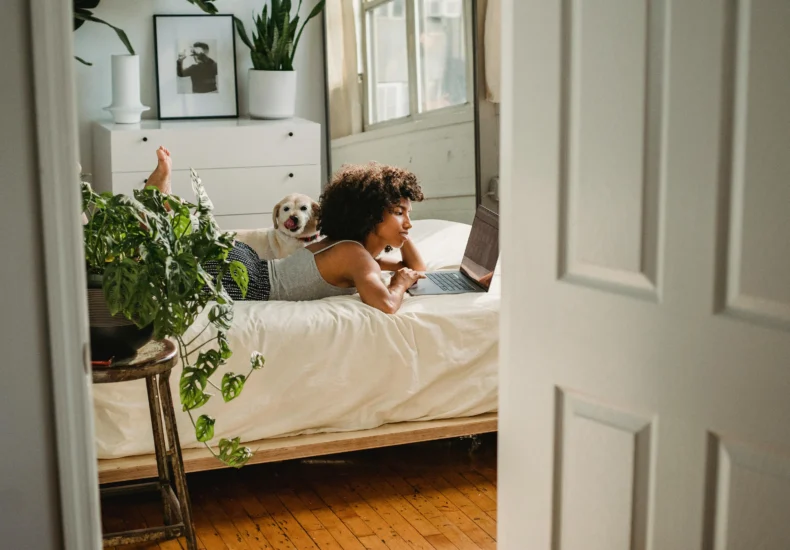 Side view of young ethnic female with netbook leaning on hand while resting on bed with dog at home