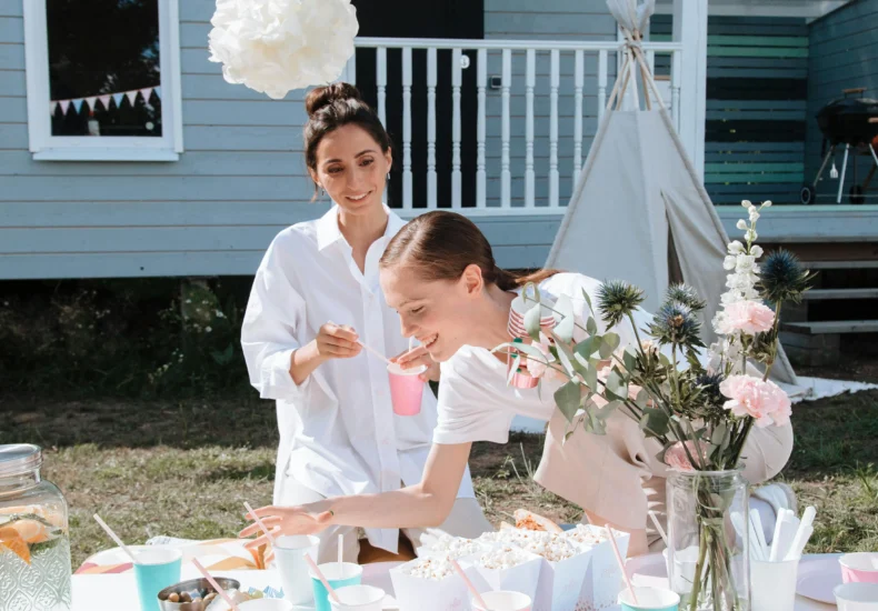 Two women enjoying a backyard party with food and floral decorations on a sunny day.