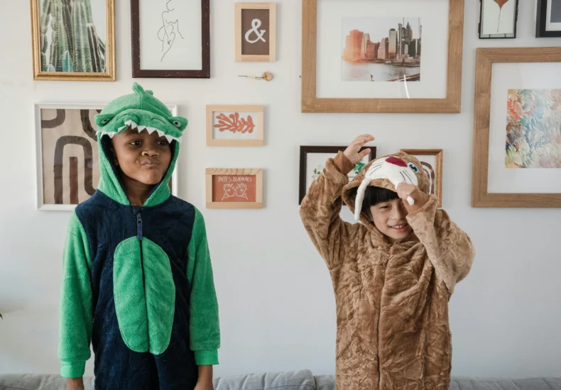 Two kids in dinosaur and dog onesies standing by a decorated wall at home.