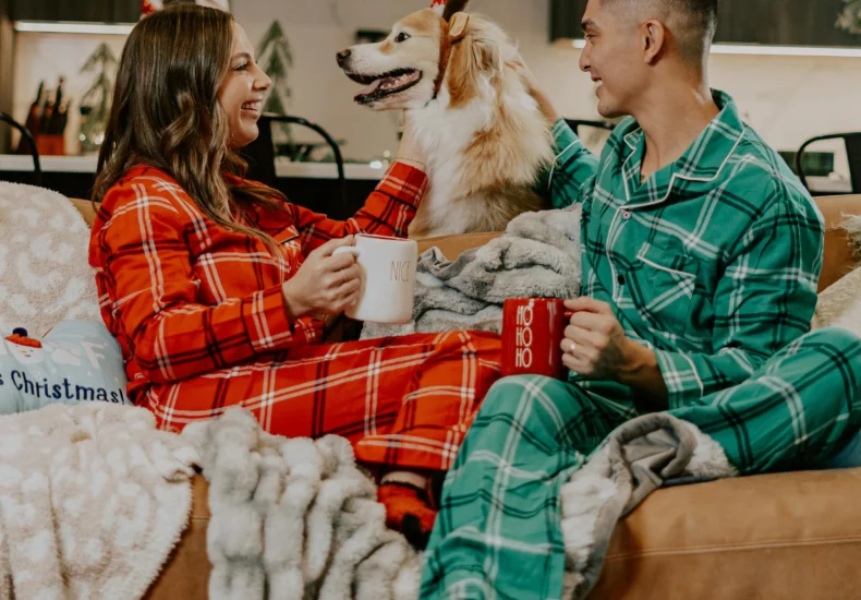 Cozy couple in plaid onesies sharing coffee with their dog on Christmas morning.