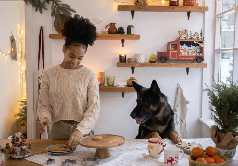 A joyful woman baking cookies with her German Shepherd in a festively decorated kitchen.