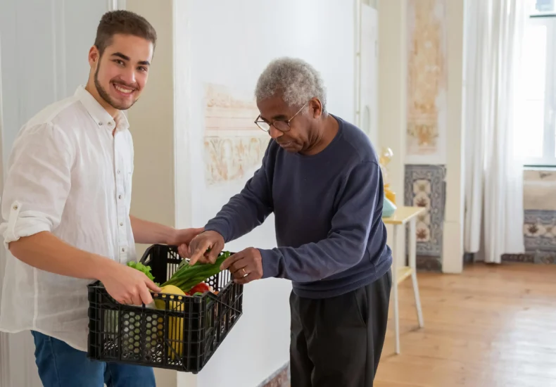 A young volunteer offers support by delivering fresh vegetables to an elderly gentleman indoors.