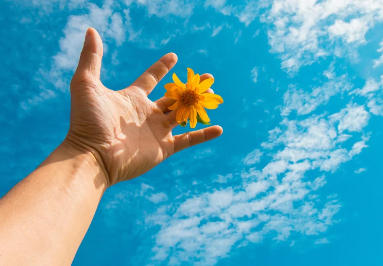 Hand holding a bright yellow flower against a textured blue sky, evoking a sense of freedom and nature.