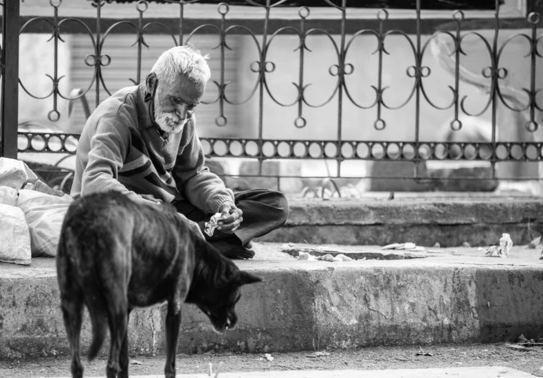 A heartwarming scene of an elderly man feeding a stray dog on a street in India, captured in monochrome.